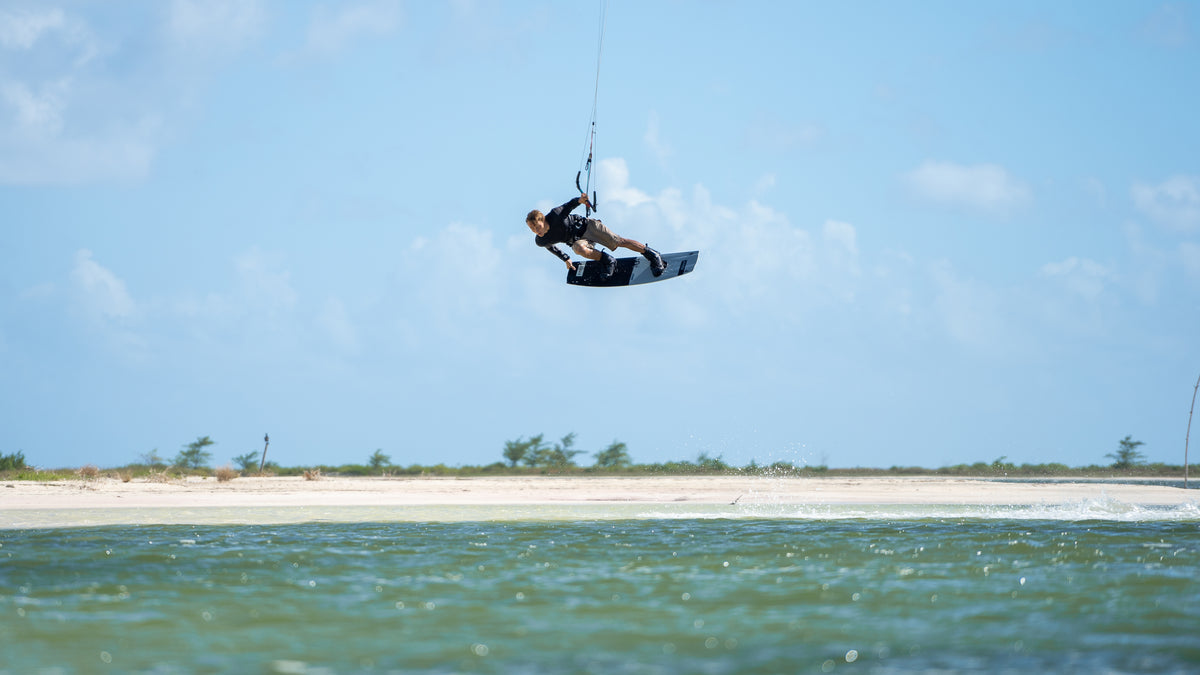 A person wakeboarding or kitesurfing in mid-air over shallow green-blue water, with a sandy shoreline and sparse trees under a bright blue sky in the background.